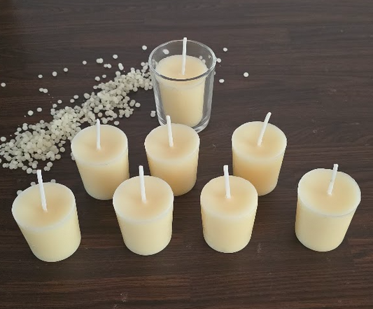 Six small cream-colored pillar candles with white wicks are arranged on a dark wooden table, with one larger candle in a clear glass holder behind them and scattered white wax beads on the left.