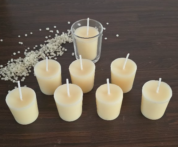 Six small cream-colored pillar candles with white wicks are arranged on a dark wooden table, with one larger candle in a clear glass holder behind them and scattered white wax beads on the left.